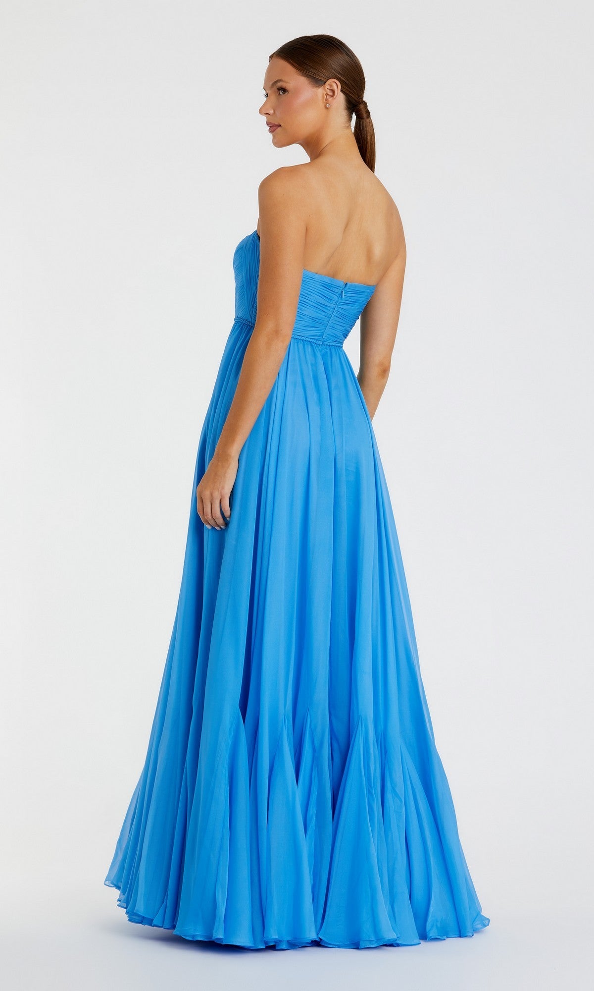 A woman stands against a plain white background, facing slightly away with her hair tied back, wearing the Mac Duggal 68847—a strapless blue chiffon gown featuring an elegant A-line skirt that exudes effortless sophistication.